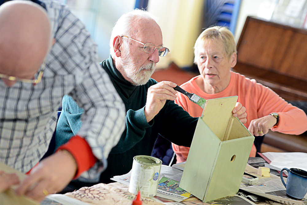 This photo shows the men's group making bird boxes. A man is seated at a table painting a bird box with a sighted helper, who is a lady helping him. Beside him is another man who is standing and working on another birdbox.
