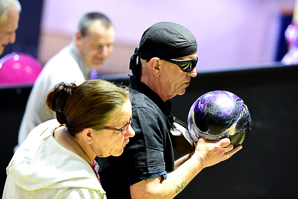 This photo shows a man with a bowling ball in his hand ready to throw, he is wearing a bandana and dark glasses. Beside him is a woman who is sighted helping him into position before he bowls the ball. They are in a ten pin bowling alley.