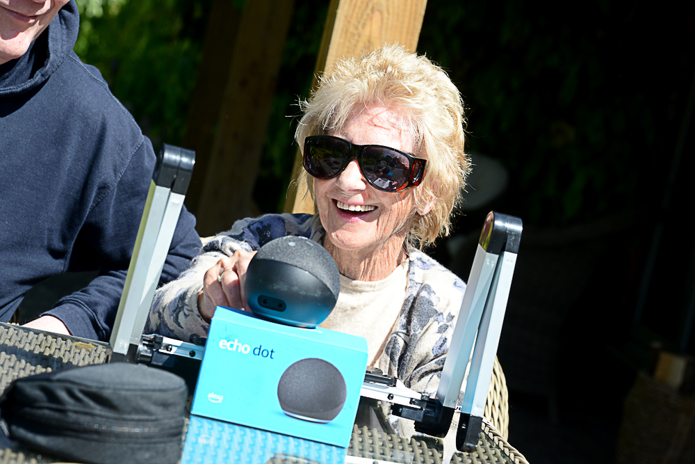 A smiling woman wearing dark glasses sat outside at a table with various gadgets in front of her.