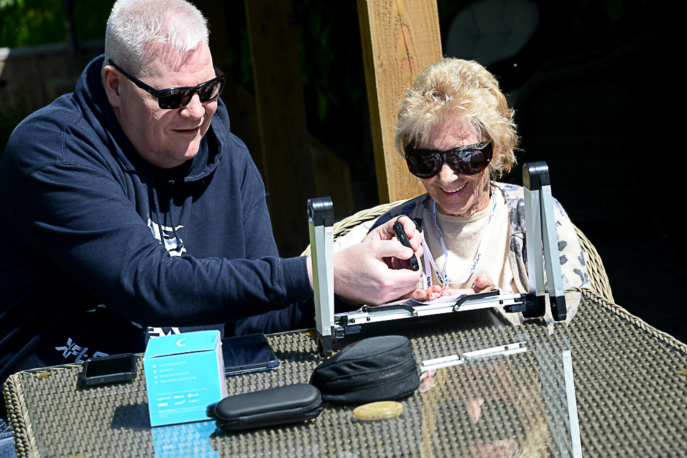 A man and a woman sat outdoors at a table in the sunshine enjoying looking at gadgets on the table in front of them, both people are wearing UV Sheilds. There is an ipad, mobile phone, Echo Dot Alexa device, glasses in cases and a cradle to elevate the magnifier.