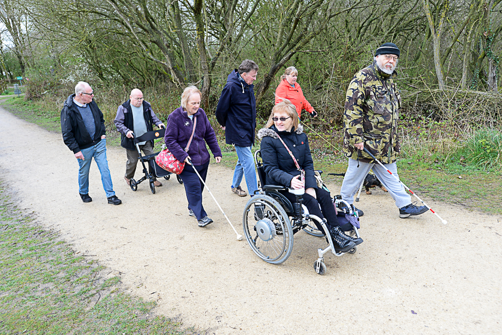 Photo shows the walking group with members walking along a path. There is a wheelchair user, someone using a white stick and a mix of men and women in the group.